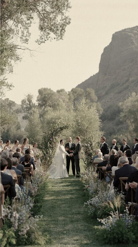 Romantic outdoor wedding ceremony set against a dramatic mountain landscape, with a floral-lined aisle, seated guests, and the bride and groom exchanging vows beneath a greenery arch.